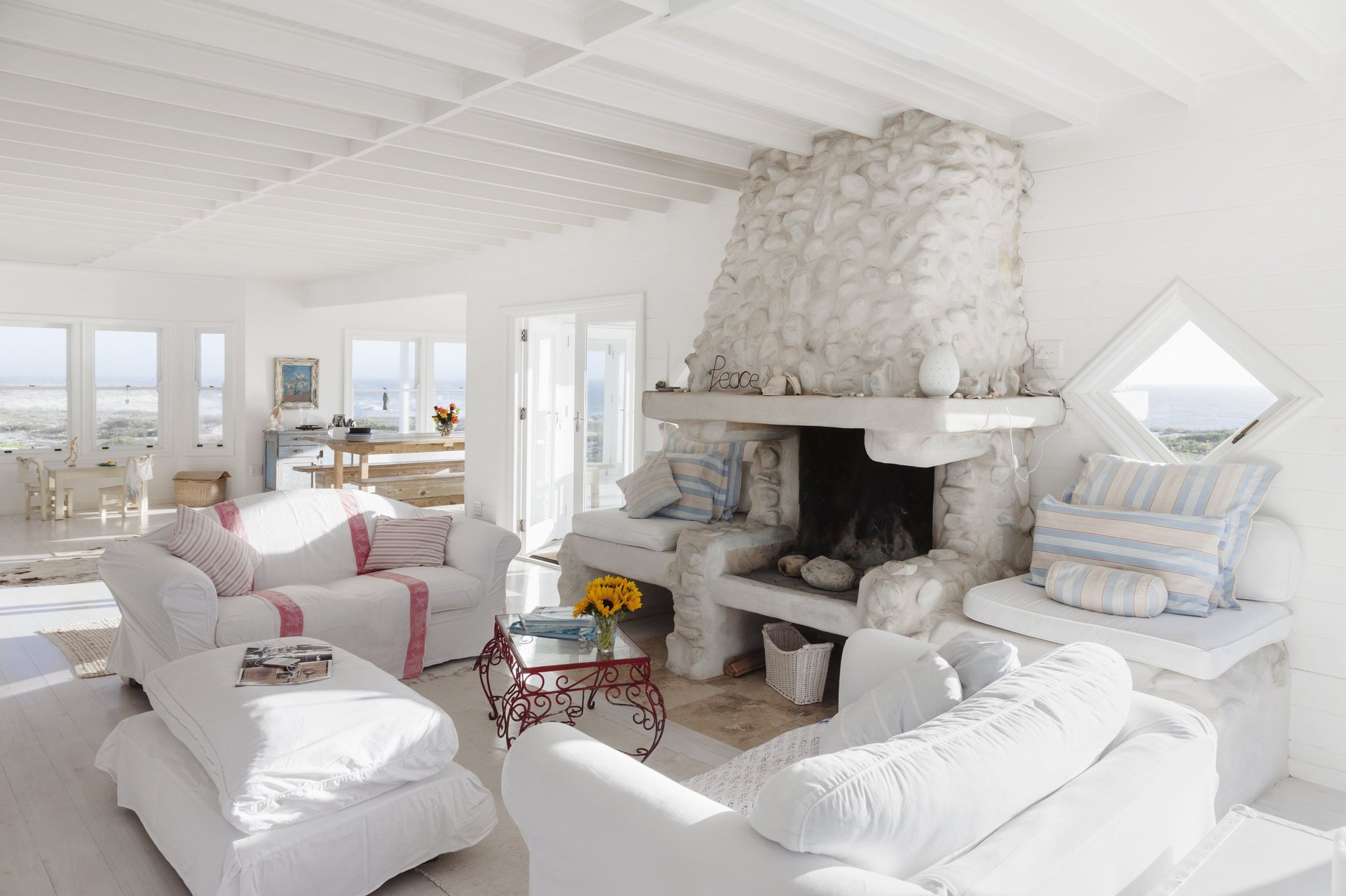 Bright living room with white sofas, a rustic stone fireplace, and large windows overlooking a coastal view