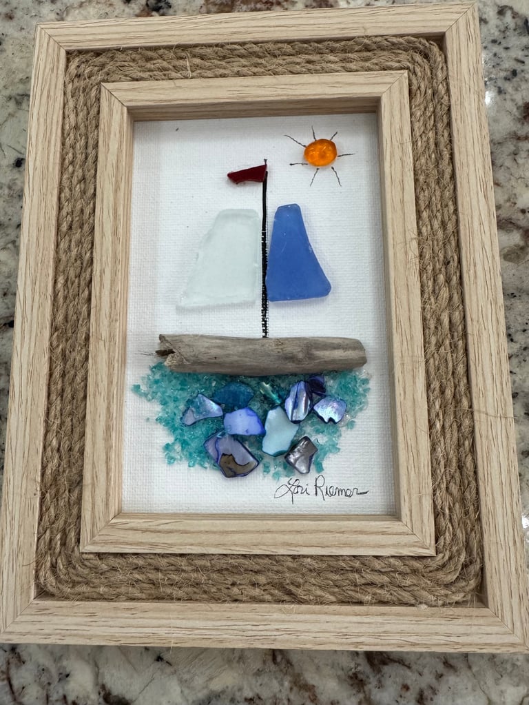 Framed coastal art made with sea glass, driftwood, and shells depicting a sailboat with red flag and sun