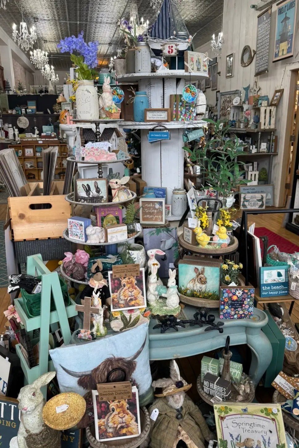 Cluttered retail display with tiered shelving, flowers, home décor items, and figurines in a shop interior