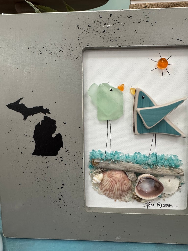 Handmade artwork with a concrete-textured frame containing a beach-themed collage with sea glass birds, shells, driftwood, and a Michigan silhouette on the left panel