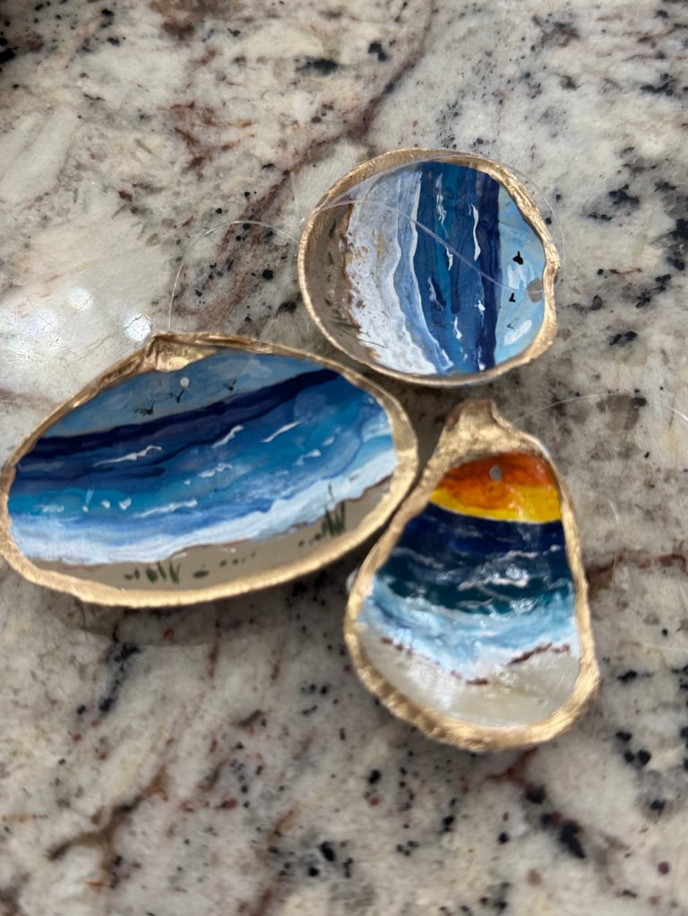 Three hand-painted seashell art pieces displaying ocean landscapes with blue water and golden sand on a speckled stone surface.
