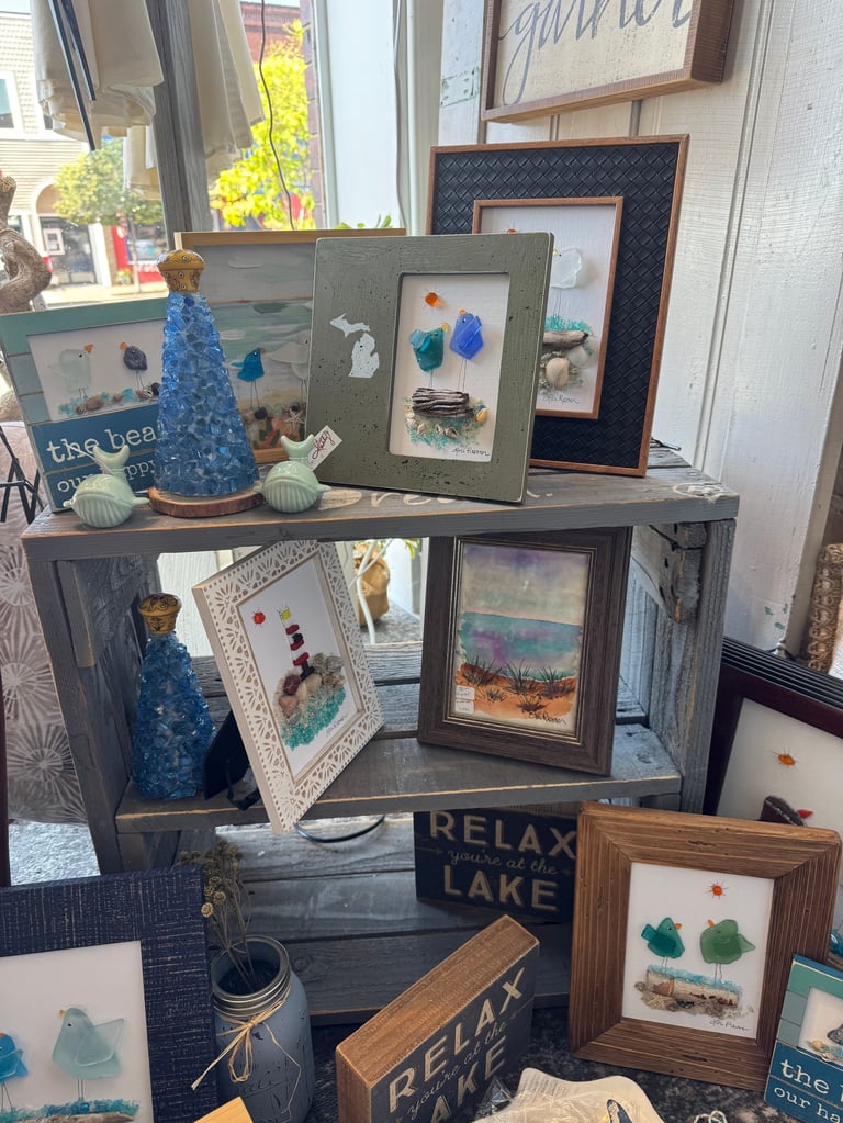 Retail display shelf showcasing framed seashell and coastal artwork, decorative bottles, and beachthemed signs and ornaments