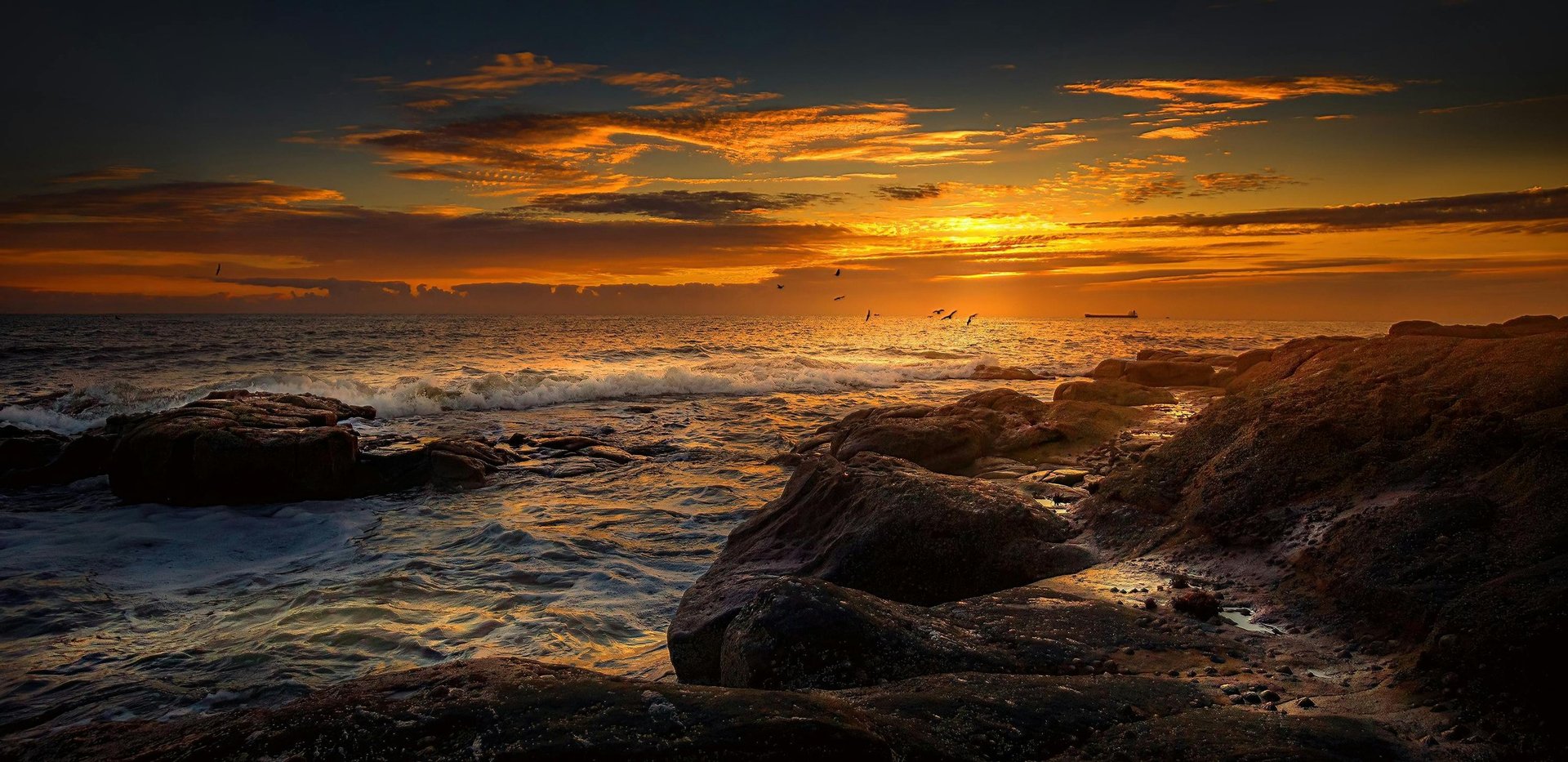 Golden sunset over ocean with rocky coastline in foreground and dramatic clouds in dark sky