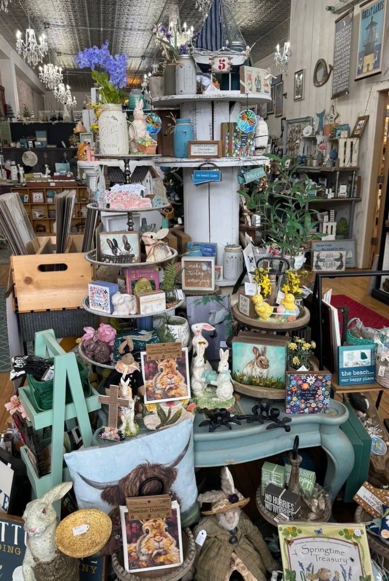 Cluttered vintage-style shop display with tiered shelving, flowers, decorative items, and antique merchandise arranged throughout