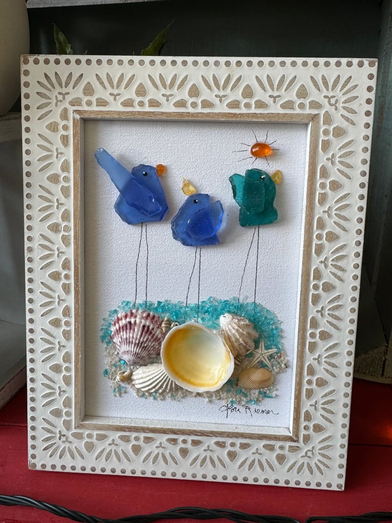 Framed mixed media art featuring three blue glass birds on thin stems above shells and beads on white fabric with decorative border