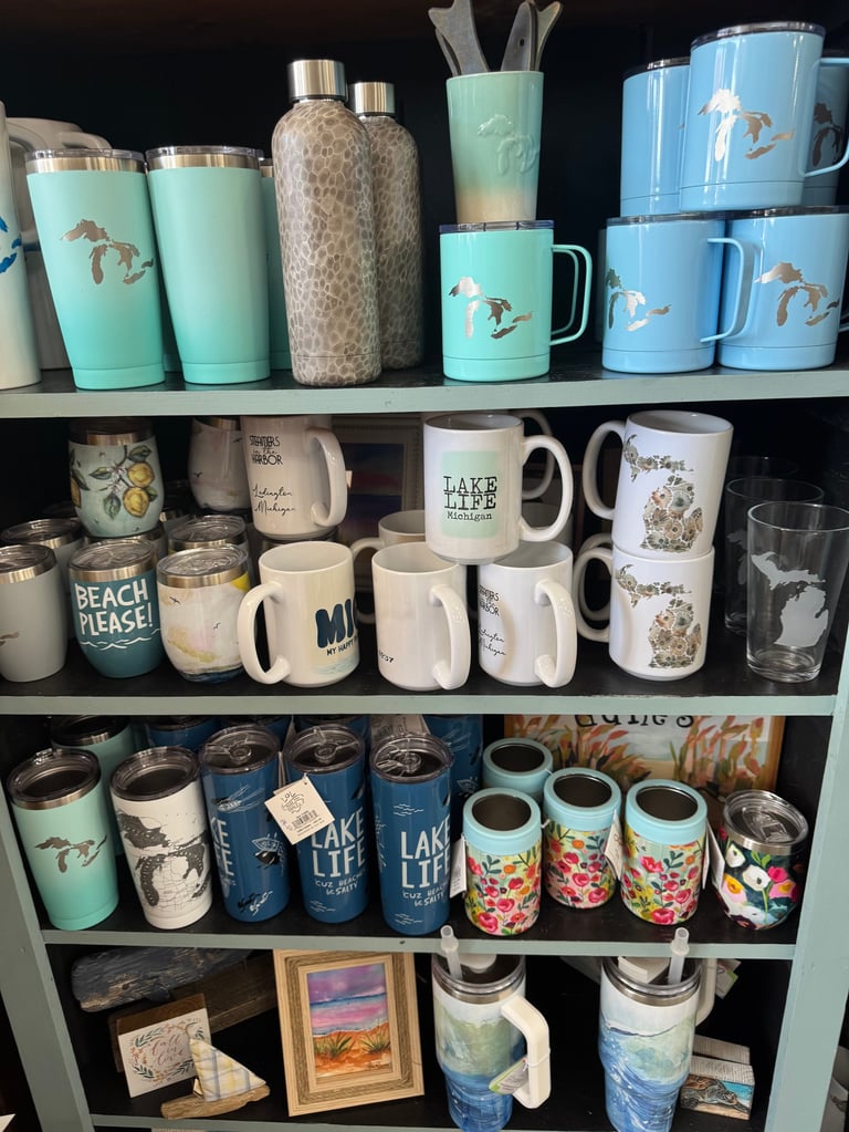 Organized shelving unit displaying a collection of insulated tumblers, mugs, and drinkware with lake-themed designs in turquoise, white, and blue.
