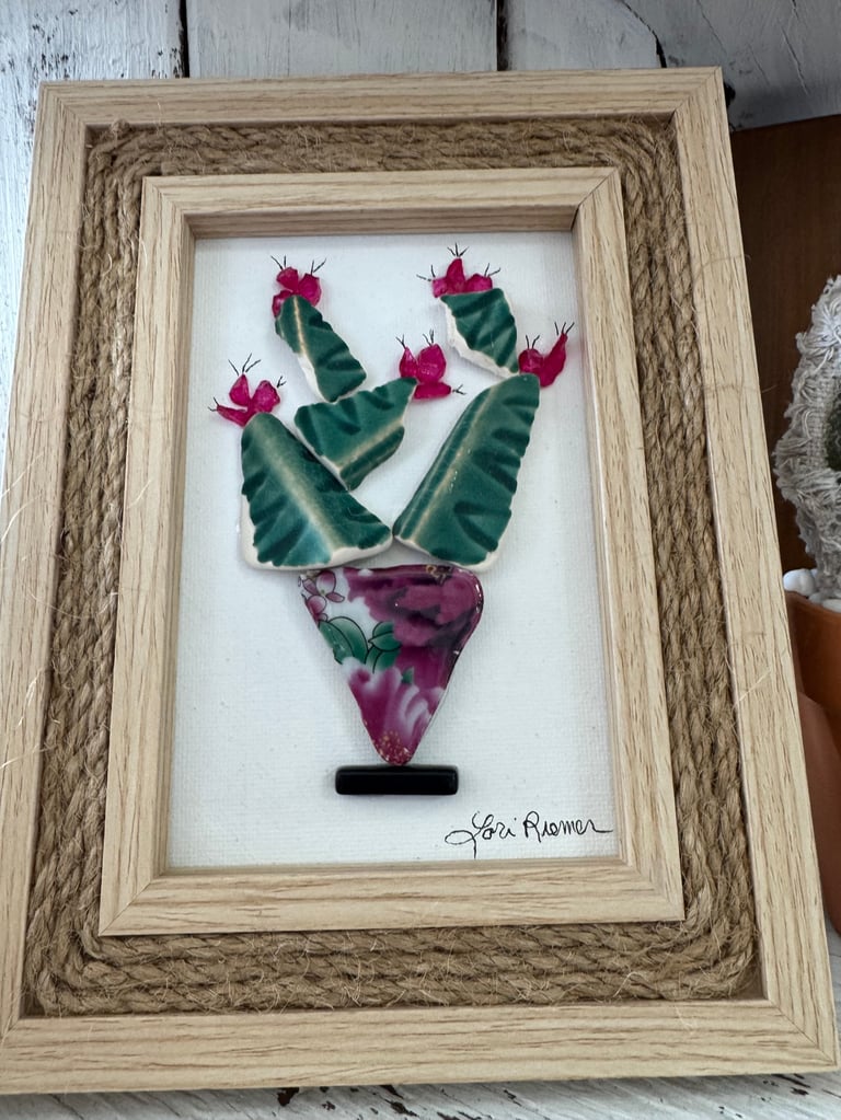 Framed botanical art featuring green cacti with hot pink flowers arranged in a V-shape above a purple succulent base, signed by artist