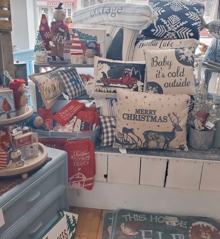 Retail display window featuring Christmas home decor with pillows, reindeer sign, vintage truck, and holiday-themed decorative items in red, navy, and cream colors